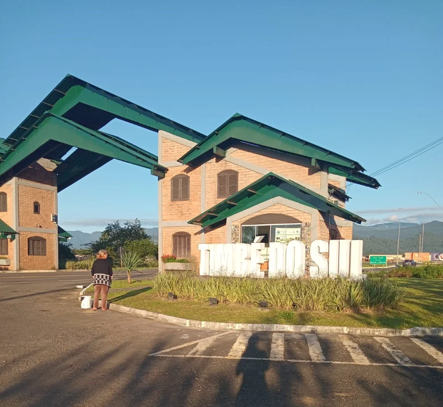 Pórtico de Timbé do Sul passa por revitalização completa Pórtico de entrada de Timbé do Sul com letreiro branco em destaque durante serviço de revitalização. A imagem mostra a estrutura arquitetônica com telhado verde, paredes de tijolos e uma colaboradora da Secretaria de Obras realizando a manutenção e pintura do local sob céu azul.