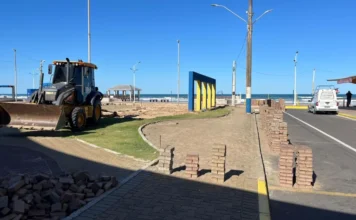 Praça Central de Arroio do Silva passa por revitalização Vista geral das obras de infraestrutura na Praça Central de Balneário Arroio do Silva, mostrando um trator John Deere amarelo estacionado, pilhas de paver organizadas ao longo da via para o novo estacionamento oblíquo, e estruturas azuis e amarelas na orla da praia sob céu limpo.