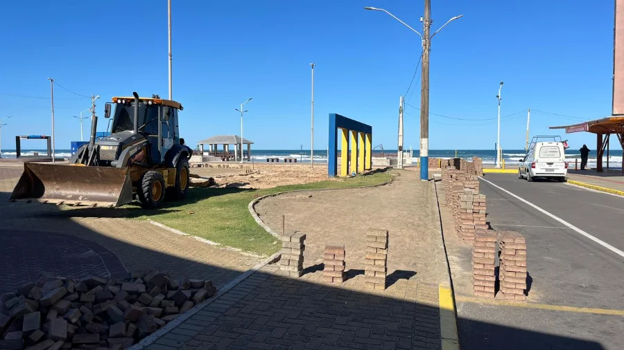Vista geral das obras de infraestrutura na Praça Central de Balneário Arroio do Silva, mostrando um trator John Deere amarelo estacionado, pilhas de paver organizadas ao longo da via para o novo estacionamento oblíquo, e estruturas azuis e amarelas na orla da praia sob céu limpo.