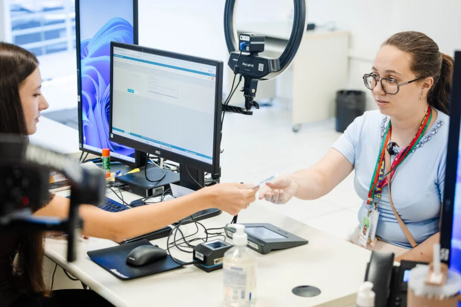 Atendimento de jovem eleitora em cartório do TRE-SC para alistamento eleitoral. Funcionária entrega cartão de título de eleitor em mesa com equipamento de biometria. Foto para matéria sobre jovens eleitores em Santa Catarina.
