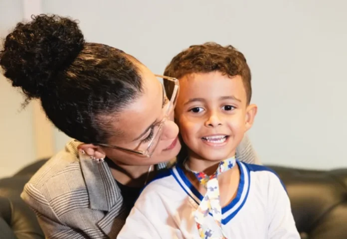 SC amplia leis e auxílio financeiro para mães atípicas Nadynne Chagas abraça o filho Matheus, uma criança com autismo nível 3 que utiliza um cordão de quebra-cabeça, simbolizando a rede de apoio e as políticas para mães atípicas em Santa Catarina.