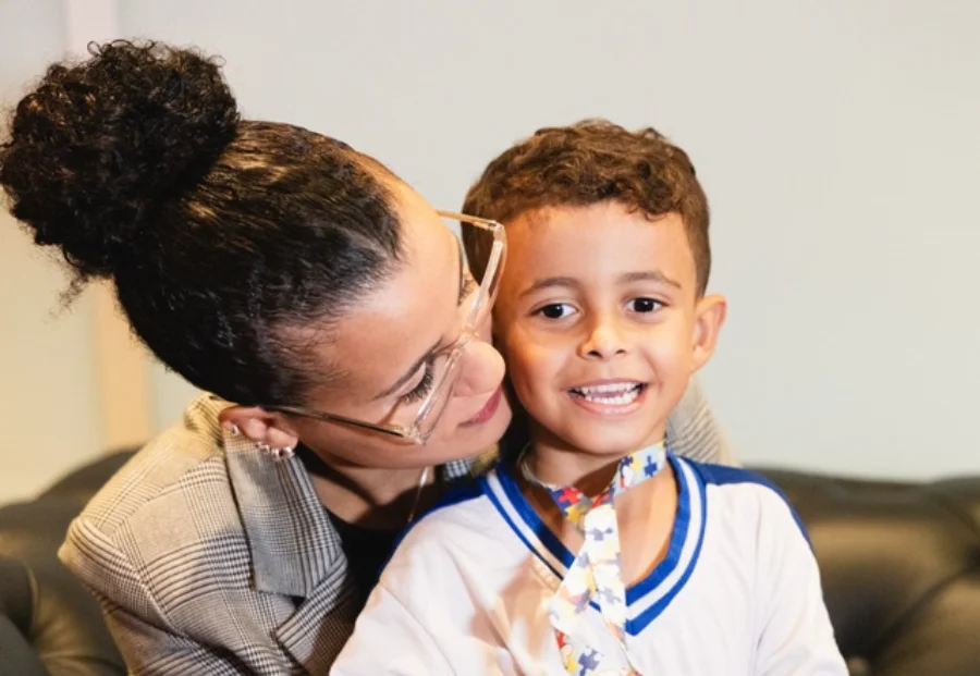 Nadynne Chagas abraça o filho Matheus, uma criança com autismo nível 3 que utiliza um cordão de quebra-cabeça, simbolizando a rede de apoio e as políticas para mães atípicas em Santa Catarina.