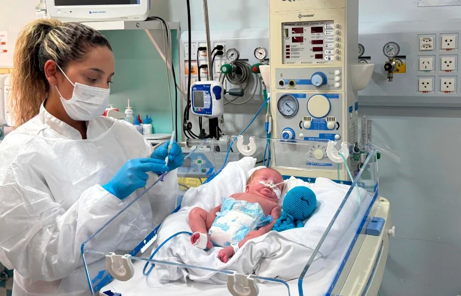 Profissional de saúde com máscara e luvas prepara o anticorpo monoclonal nirsevimabe em seringa ao lado de bebê prematuro em incubadora hospitalar em Santa Catarina. A imagem ilustra a prevenção contra o Vírus Sincicial Respiratório (VSR) em recém-nascidos vulneráveis.