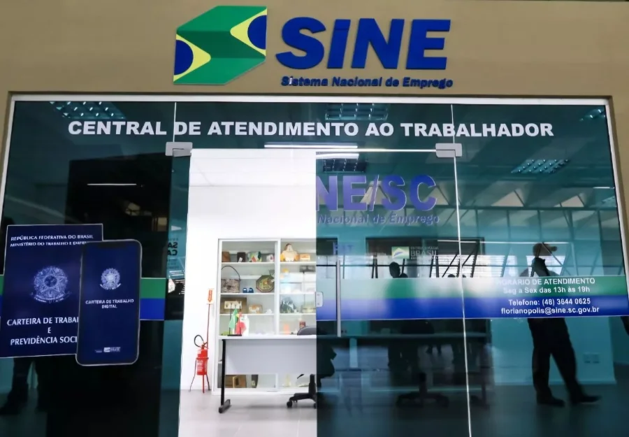 Fachada de vidro da entrada de uma unidade do Sine em Santa Catarina, exibindo a logomarca do Sistema Nacional de Emprego e a frase Central de Atendimento ao Trabalhador. Ao lado da porta, há um adesivo ilustrativo da Carteira de Trabalho Digital.
