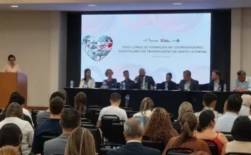 SC promove curso para coordenadores de transplante Mesa de abertura do XXXIII Curso de Formação de Coordenadores Hospitalares de Transplante de Santa Catarina, em Florianópolis. Painel com autoridades e palestrante falando ao microfone diante de uma audiência sentada. Tela de fundo exibe logos da SC Transplantes e do Governo do Estado, além do título do evento.