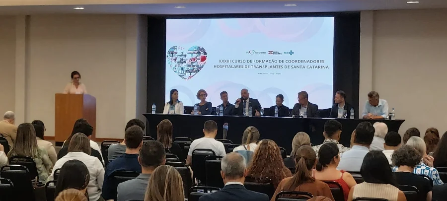 SC promove curso para coordenadores de transplante Mesa de abertura do XXXIII Curso de Formação de Coordenadores Hospitalares de Transplante de Santa Catarina, em Florianópolis. Painel com autoridades e palestrante falando ao microfone diante de uma audiência sentada. Tela de fundo exibe logos da SC Transplantes e do Governo do Estado, além do título do evento.