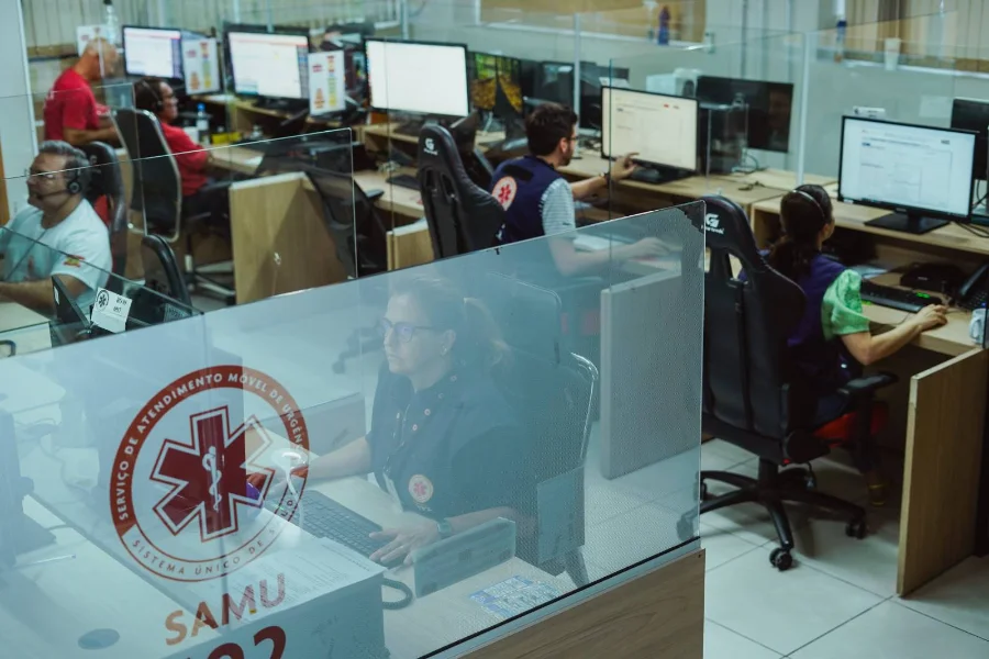 Profissionais trabalham na Central de Regulação das Urgências do Samu em Santa Catarina, operando computadores e headsets para o atendimento de chamados do 192 em ambiente com divisórias de vidro.