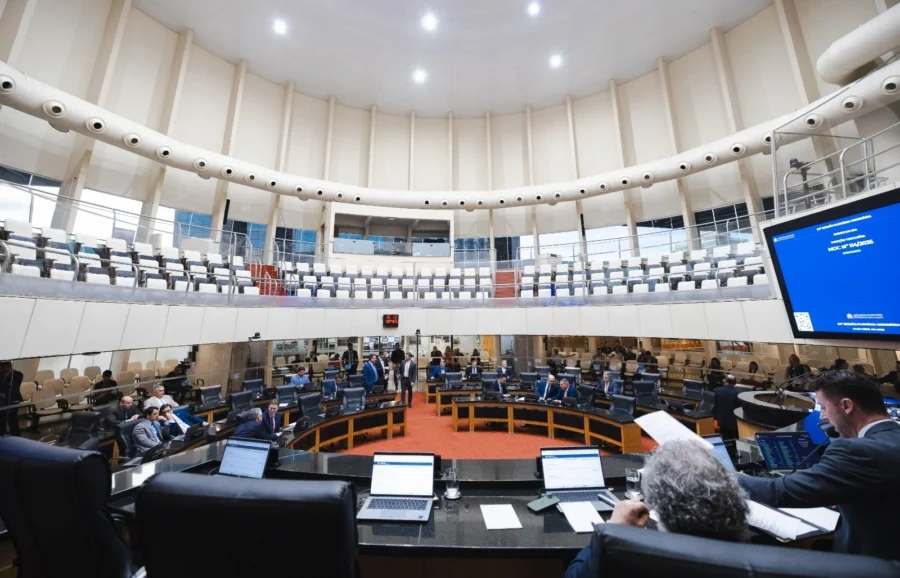 Vista em grande angular do plenário circular da Assembleia Legislativa de Santa Catarina (Alesc) durante uma sessão parlamentar movimentada, mostrando deputados sentados em suas bancadas, servidores com laptops em primeiro plano e uma grande tela exibindo informações sobre a 38ª Sessão Plenária Ordinária.