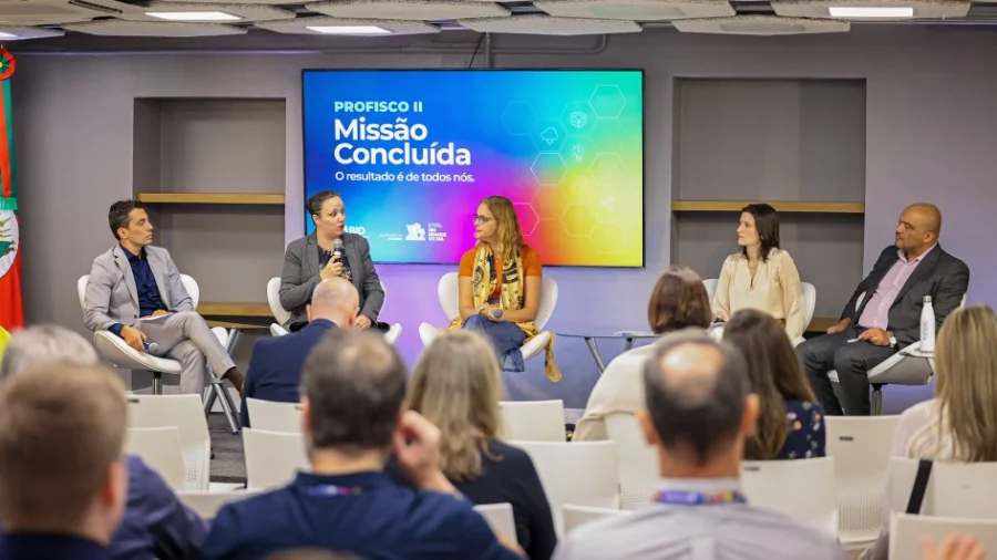 Sefaz-RS encerra Profisco II com avanços tecnológicos Fotografia de oradores em um painel de encerramento do programa de modernização fiscal Profisco II na Sefaz RS. O ecrã de fundo mostra 'Missão Concluída' e os logótipos do BID e do Governo do Rio Grande do Sul. Plateia desfocada em primeiro plano. Foco em gestão tributária e eficiência fiscal.
