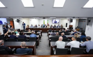 Setor de supermercados ganha frente parlamentar na ALRS Fotografia de ângulo alto do Plenarinho da Assembleia Legislativa do Rio Grande do Sul (ALRS), lotado durante a instalação da Frente Parlamentar em Defesa do Setor Supermercadista Gaúcho. A imagem captura a mesa diretora composta por deputados e líderes do varejo, incluindo o presidente da frente, deputado Guilherme Pasin, e o presidente da AGAS, Lindonor Peruzzo Júnior. Oradores se dirigem ao público e autoridades.