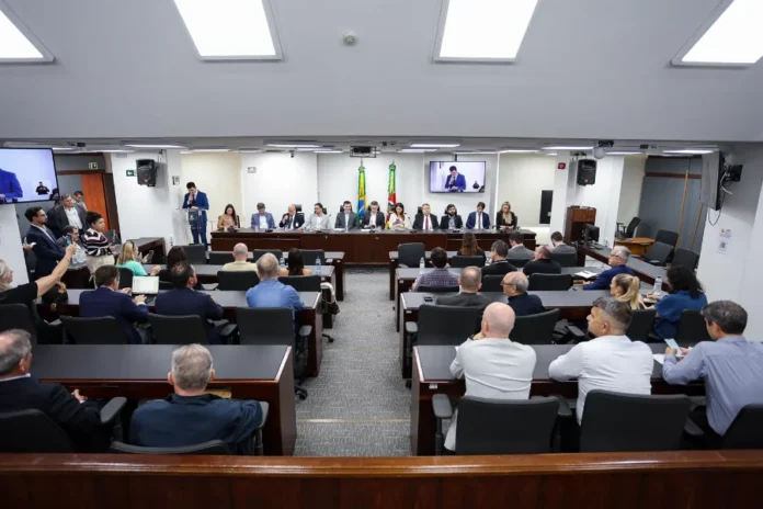 Setor de supermercados ganha frente parlamentar na ALRS Fotografia de ângulo alto do Plenarinho da Assembleia Legislativa do Rio Grande do Sul (ALRS), lotado durante a instalação da Frente Parlamentar em Defesa do Setor Supermercadista Gaúcho. A imagem captura a mesa diretora composta por deputados e líderes do varejo, incluindo o presidente da frente, deputado Guilherme Pasin, e o presidente da AGAS, Lindonor Peruzzo Júnior. Oradores se dirigem ao público e autoridades.