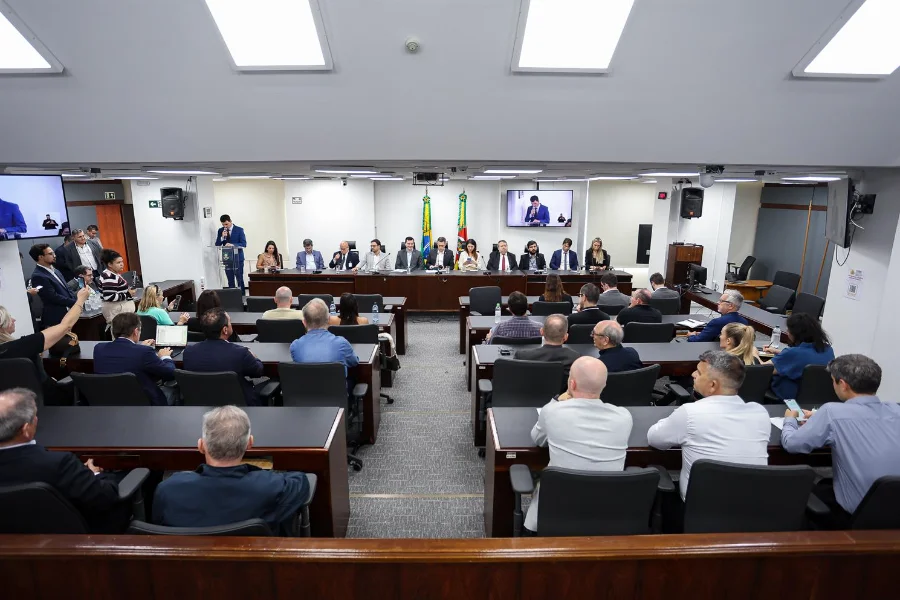 Fotografia de ângulo alto do Plenarinho da Assembleia Legislativa do Rio Grande do Sul (ALRS), lotado durante a instalação da Frente Parlamentar em Defesa do Setor Supermercadista Gaúcho. A imagem captura a mesa diretora composta por deputados e líderes do varejo, incluindo o presidente da frente, deputado Guilherme Pasin, e o presidente da AGAS, Lindonor Peruzzo Júnior. Oradores se dirigem ao público e autoridades.