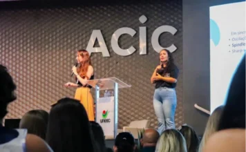 Simpósio debate autismo na vida adulta e envelhecimento Foto de uma palestrante em um púlpito com o logotipo da UNESC, em um palco no auditório da ACIC durante o Simpósio LAND sobre autismo em Criciúma. À direita, uma intérprete de Libras realiza a tradução para a plateia visível em primeiro plano. O letreiro da ACIC está na parede ao fundo.