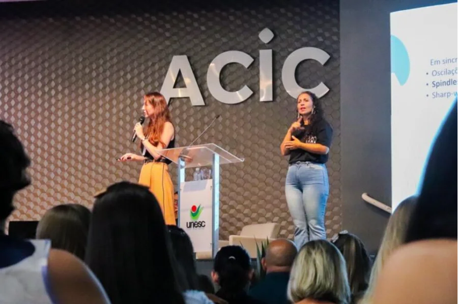 Foto de uma palestrante em um púlpito com o logotipo da UNESC, em um palco no auditório da ACIC durante o Simpósio LAND sobre autismo em Criciúma. À direita, uma intérprete de Libras realiza a tradução para a plateia visível em primeiro plano. O letreiro da ACIC está na parede ao fundo.
