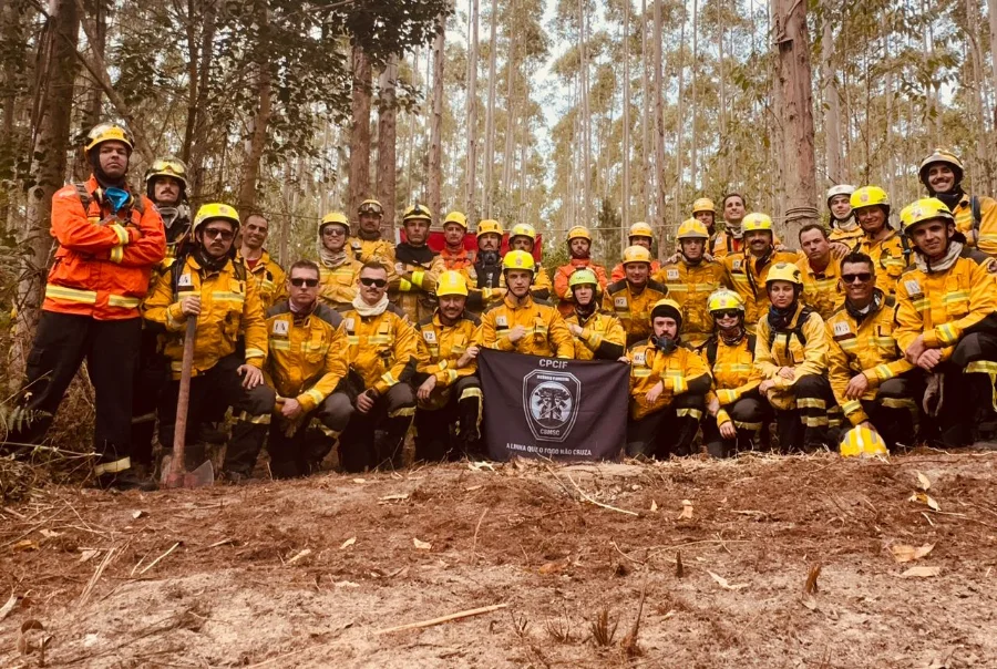 Bombeiros militares de Santa Catarina em uniforme de combate posam para foto oficial em área de mata em Sombrio, segurando bandeira do Curso de Prevenção e Combate a Incêndio Florestal (CPCIF).