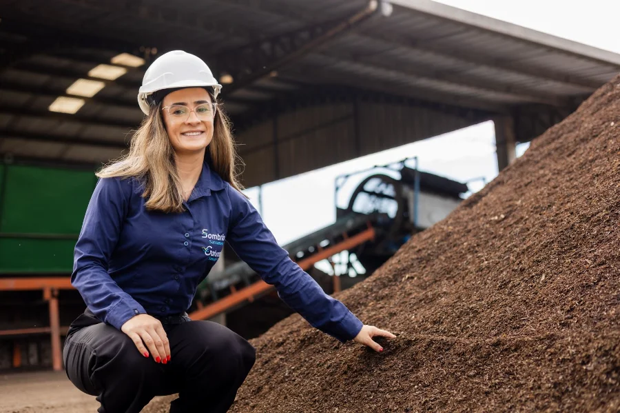 Profissional da Sombrio Saneamento, Camila Lima, usando EPI e agachada ao lado de uma pilha de fertilizante orgânico produzido a partir de lodo de esgoto tratado na instalação da Composul Compostagem, exemplificando o reaproveitamento sustentável de resíduos.