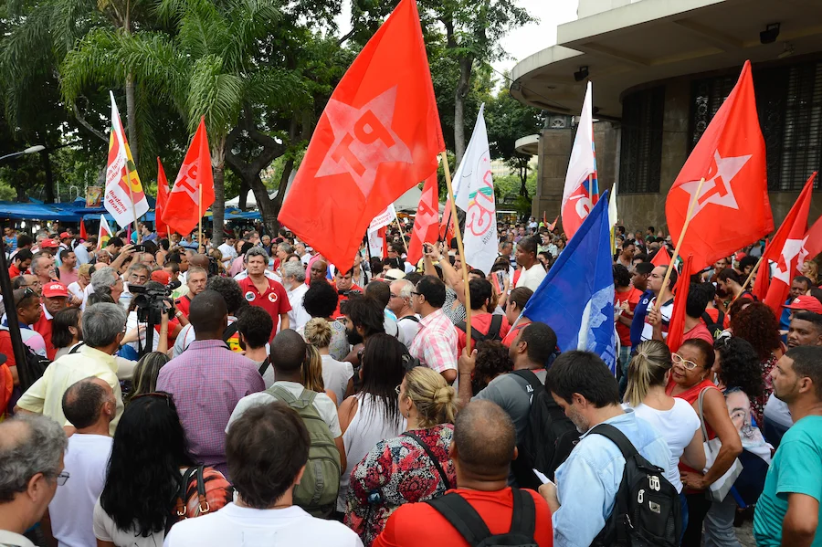 Manifestação política com militantes e apoiadores do PT segurando bandeiras vermelhas da sigla em praça pública, representando a base partidária que aguarda as diretrizes do 8º Congresso.