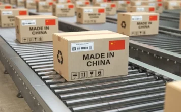 Taxa das blusinhas preserva 135 mil empregos no Brasil Uma esteira de remessa industrial carregada com caixas de papelão. A caixa em destaque tem a etiqueta "MADE IN CHINA", uma bandeira da China e símbolos de envio internacional, ilustrando o fluxo de mercadorias importadas no contexto do comércio exterior e a nova taxação de blusinhas.