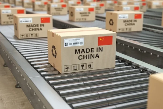 Taxa das blusinhas preserva 135 mil empregos no Brasil Uma esteira de remessa industrial carregada com caixas de papelão. A caixa em destaque tem a etiqueta "MADE IN CHINA", uma bandeira da China e símbolos de envio internacional, ilustrando o fluxo de mercadorias importadas no contexto do comércio exterior e a nova taxação de blusinhas.