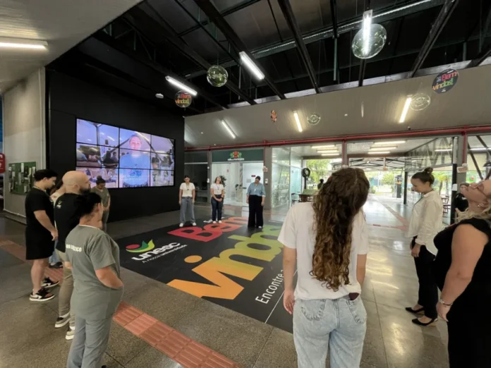 Unesc amplia ações de saúde física para seus funcionários "Grupo de colaboradores da Unesc realiza exercícios de ginástica laboral e alongamento postural no saguão da universidade, focando na saúde e bem-estar do trabalhador. O logo da Unesc está visível no adesivo de chão."