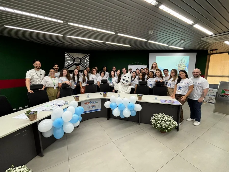 Grupo de acadêmicos de saúde da Unesc e profissionais reunidos no Paço Municipal de Criciúma para o lançamento do projeto VER-SUS/Brasil. A imagem mostra cerca de 30 pessoas com camisetas brancas do projeto, posicionadas atrás de uma bancada com balões azuis e brancos e a presença do mascote Zé Gotinha ao centro.