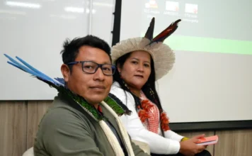 Unesc inicia 8ª Semana Indígena com foco em resistência Lideranças indígenas Kerexu Yxapyry e Fabiano Alves (Karai) sentados em mesa de debate durante a abertura da 8ª Semana Indígena na Unesc, em Criciúma. Kerexu usa cocar de penas claras e Fabiano utiliza adorno de penas coloridas no pescoço.