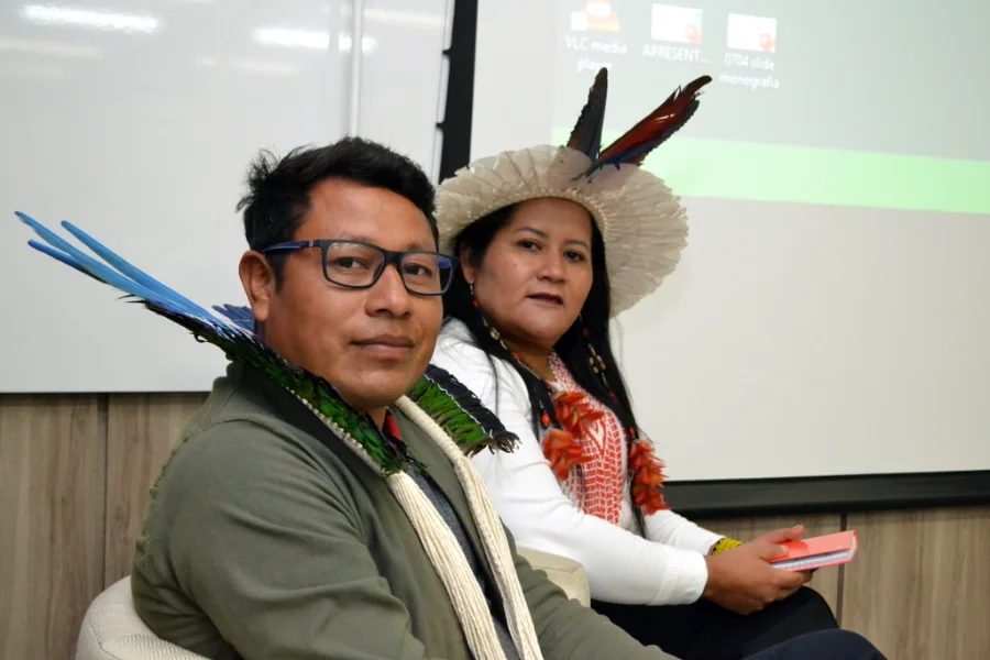 Lideranças indígenas Kerexu Yxapyry e Fabiano Alves (Karai) sentados em mesa de debate durante a abertura da 8ª Semana Indígena na Unesc, em Criciúma. Kerexu usa cocar de penas claras e Fabiano utiliza adorno de penas coloridas no pescoço.