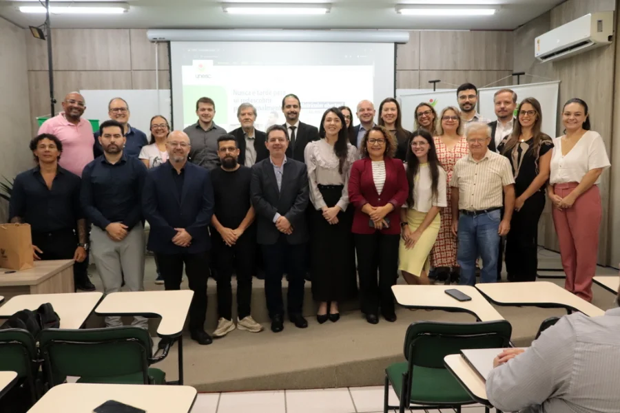 Grupo de professores e alunos da primeira turma do Doutorado em Direito da Unesc reunidos em sala de aula para a aula magna em Criciúma. Ao fundo, tela de projeção e banners da universidade. Foto com foco na formação jurídica e Direitos Humanos.
