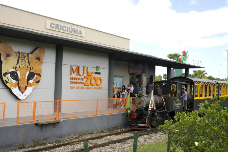 Fachada do Museu de Zoologia da Unesc (Muzoo) no Parque das Nações em Criciúma, exibindo pintura de um gato-maracajá na parede e o trem turístico Terezinha parado na estação em frente ao museu.