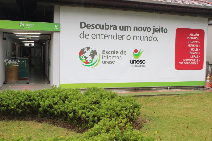 Fachada da Escola de Idiomas da Unesc com painel informativo listando cursos como italiano, inglês e espanhol. A imagem destaca a entrada da unidade onde será realizado o curso de italiano para profissionais da saúde.