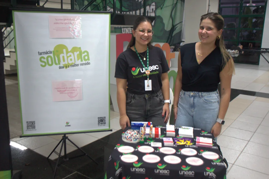 Acadêmicas de Farmácia da Unesc, Liandra Miotelli e Mariane Raupp, sorriem ao lado de banner da Farmácia Solidária e mesa com amostras de anticoncepcionais durante ação de conscientização sobre saúde feminina no Bloco S da universidade.