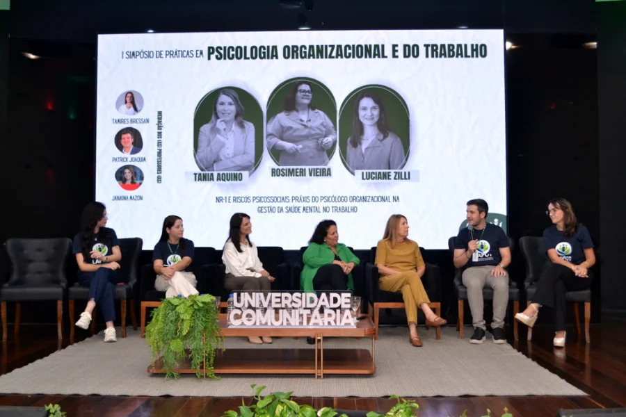Palestrantes e acadêmicos em mesa-redonda durante o 1º Simpósio de Psicologia Organizacional e do Trabalho na Unesc, debatendo a gestão da saúde mental no trabalho e a aplicação da NR-1 em auditório da universidade.