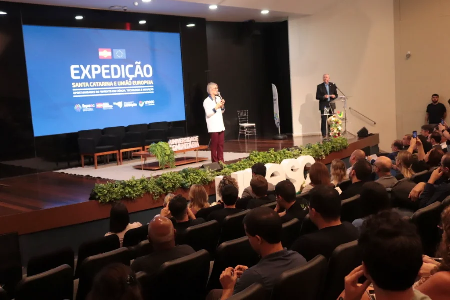 Reitora em exercício da Unesc, Gisele Silveira Coelho Lopes, discursa no palco durante a 4ª Expedição Santa Catarina e União Europeia em Criciúma. Ao fundo, tela azul exibe o nome do evento sobre oportunidades de fomento em ciência, tecnologia e inovação para pesquisadores e empreendedores.