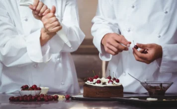 Urussanga abre inscrições para curso gratuito de confeitaria Confeiteiros com uniformes brancos decorando bolo de chocolate com chantilly e cerejas frescas, ilustrando as técnicas de produção artesanal ensinadas no curso gratuito de confeitaria em Urussanga.