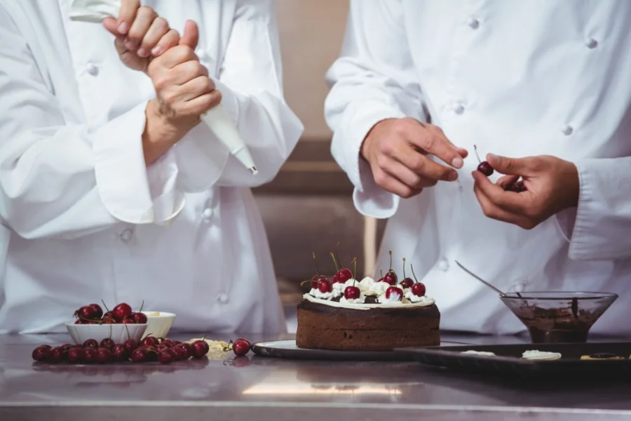Confeiteiros com uniformes brancos decorando bolo de chocolate com chantilly e cerejas frescas, ilustrando as técnicas de produção artesanal ensinadas no curso gratuito de confeitaria em Urussanga.