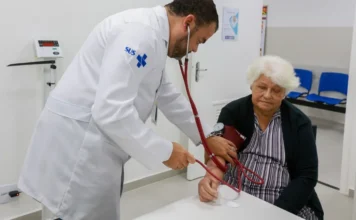 VSR avança no Brasil e acende alerta para idosos em 2026 Médico com jaleco oficial do SUS utiliza estetoscópio e esfigmomanômetro para aferir a pressão arterial de uma mulher idosa. A imagem destaca o atendimento clínico e preventivo à terceira idade, grupo de risco para complicações do vírus sincicial respiratório (VSR) e outras síndromes respiratórias no Brasil.