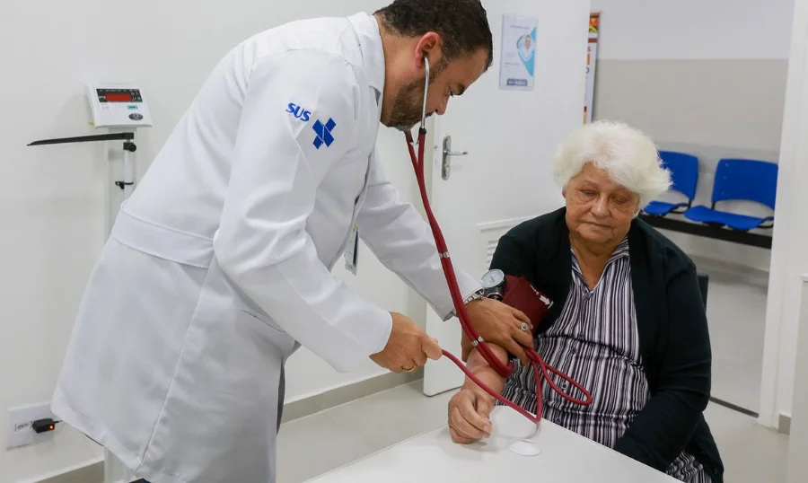 Médico com jaleco oficial do SUS utiliza estetoscópio e esfigmomanômetro para aferir a pressão arterial de uma mulher idosa. A imagem destaca o atendimento clínico e preventivo à terceira idade, grupo de risco para complicações do vírus sincicial respiratório (VSR) e outras síndromes respiratórias no Brasil.