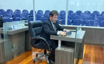 Vereadores aprovam incentivo para agricultura em Araranguá Um vereador de terno e gravata opera um pequeno painel de votação eletrônico em sua mesa na Câmara Municipal de Araranguá, cercado por fileiras de cadeiras vazias no plenário.