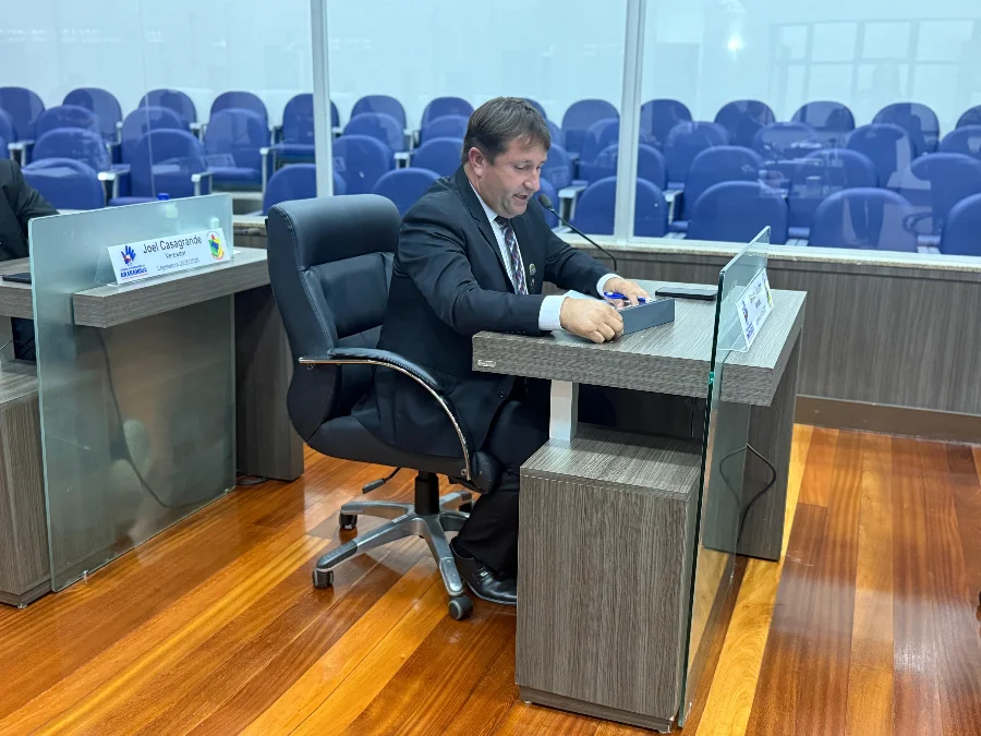 Um vereador de terno e gravata opera um pequeno painel de votação eletrônico em sua mesa na Câmara Municipal de Araranguá, cercado por fileiras de cadeiras vazias no plenário.