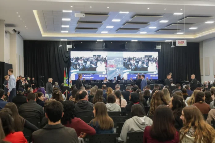 Alesc Itinerante reúne 1,5 mil pessoas em Araranguá Plenário do programa Alesc Itinerante em Araranguá com público assistindo à sessão da Assembleia Legislativa de Santa Catarina no Centro Multiuso Arlete Cristiano Salomé. Ao fundo, grande painel de LED exibe a transmissão do evento e a identidade visual do projeto.