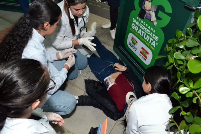Alunos de Enfermagem da Unesc treinam emergência em campus Estudantes de enfermagem da Unesc realizando atendimento de emergência simulado em vítima de trauma com colar cervical e seringa no chão durante atividade prática no campus.