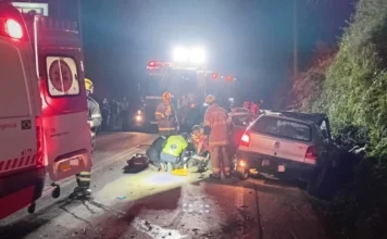 Colisão tripla deixa três feridos na SC-390 em Lauro Müller Resgate noturno na rodovia SC-390 em Lauro Müller, SC, mostrando bombeiros militares e equipe do SAMU prestando atendimento médico de emergência a uma vítima de acidente ao lado de um veículo VW Gol prateado danificado. Uma ambulância e um caminhão de bombeiros com luzes de emergência ligadas estão no local, iluminando a cena da colisão frontal tripla.