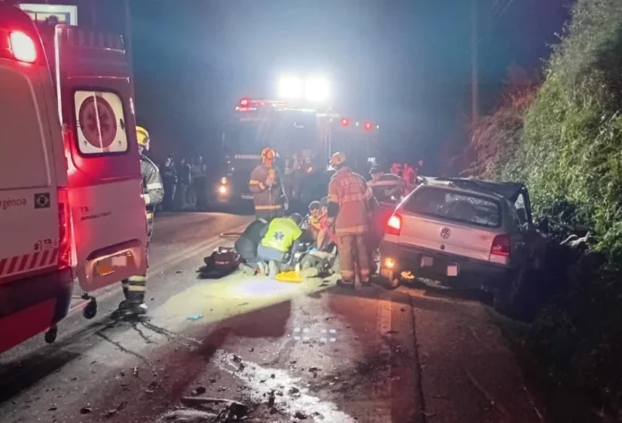 Colisão tripla deixa três feridos na SC-390 em Lauro Müller Resgate noturno na rodovia SC-390 em Lauro Müller, SC, mostrando bombeiros militares e equipe do SAMU prestando atendimento médico de emergência a uma vítima de acidente ao lado de um veículo VW Gol prateado danificado. Uma ambulância e um caminhão de bombeiros com luzes de emergência ligadas estão no local, iluminando a cena da colisão frontal tripla.