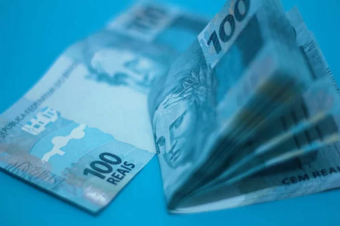 Endividamento das famílias bate recorde histórico em 2026 Cédulas de 100 reais em destaque com iluminação azulada, ilustrando a crise financeira, o endividamento recorde das famílias brasileiras e a pressão sobre a renda mensal em 2026.