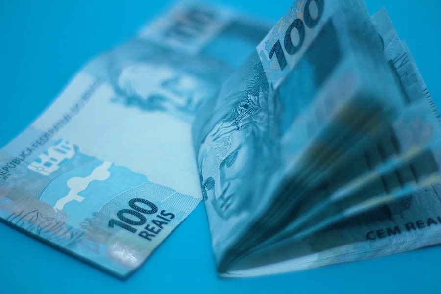 Cédulas de 100 reais em destaque com iluminação azulada, ilustrando a crise financeira, o endividamento recorde das famílias brasileiras e a pressão sobre a renda mensal em 2026.