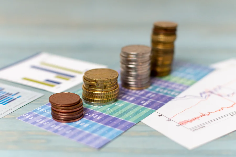 Pilhas de moedas sobre gráficos financeiros ilustram projeções do Boletim Focus para inflação, PIB e juros no Brasil.