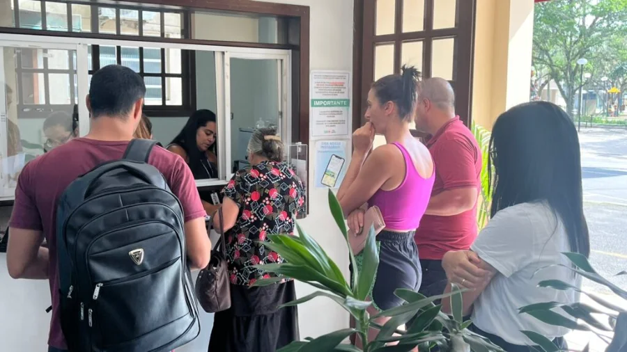 Moradores de Forquilhinha aguardam atendimento em guichê da Secretaria de Saúde para o programa Fila Zero, focado em agilizar consultas e exames especializados. A imagem mostra pessoas em fila na recepção, com cartazes informativos e uma atendente ao fundo, descrevendo o fluxo de serviços públicos para acessibilidade e SEO.