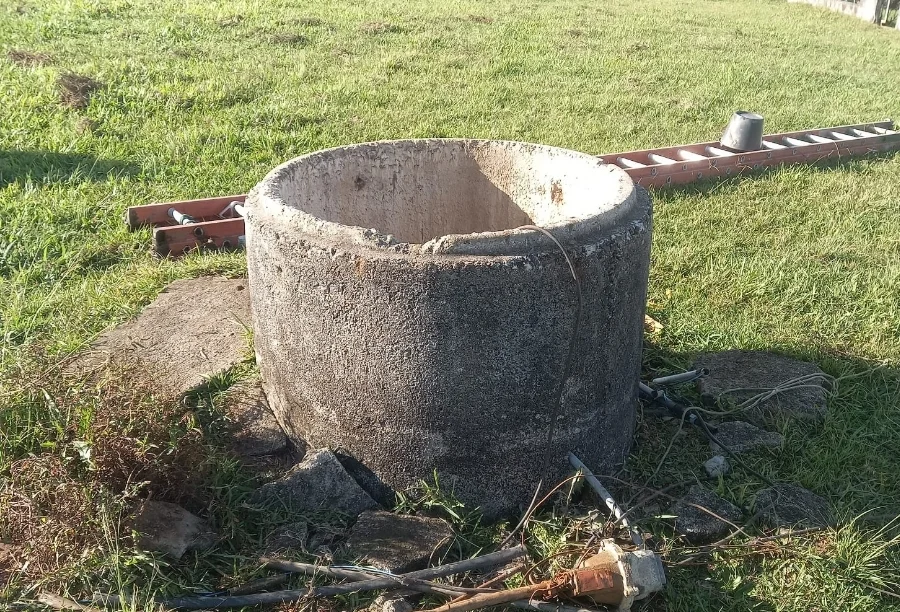 Poço de concreto em área gramada no bairro Poço 3, em Içara, local onde um homem de 58 anos foi resgatado pelo Corpo de Bombeiros após queda de uma altura de 11 metros. Ao fundo, vê-se uma escada de madeira sobre a grama.