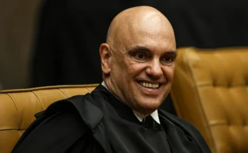 Moraes ordena prisão de empresário por bloqueios em SC Ministro do Supremo Tribunal Federal (STF), Alexandre de Moraes, sorrindo durante sessão oficial. Ele veste a toga jurídica preta e está sentado em uma poltrona de couro bege. Fotografia focada no rosto do magistrado com fundo escuro.