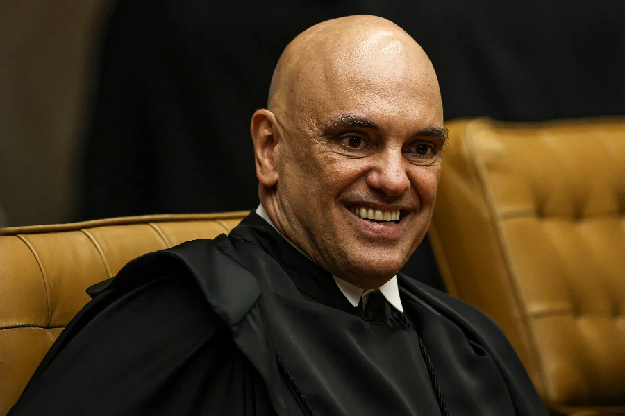 Ministro do Supremo Tribunal Federal (STF), Alexandre de Moraes, sorrindo durante sessão oficial. Ele veste a toga jurídica preta e está sentado em uma poltrona de couro bege. Fotografia focada no rosto do magistrado com fundo escuro.
